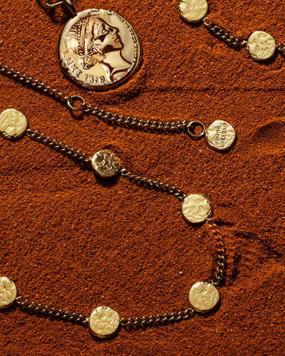 Gold Luke 15:9 coin pendant necklace and matching coin bracelets arranged on textured rust-coloured sand.