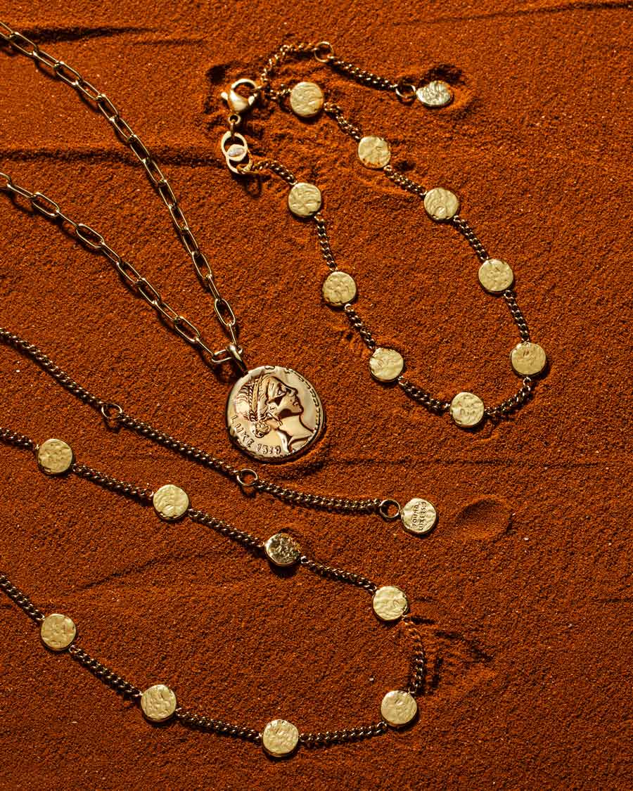 Gold Luke 15:9 coin pendant necklace and matching coin bracelet and necklace arranged on textured rust-coloured sand.