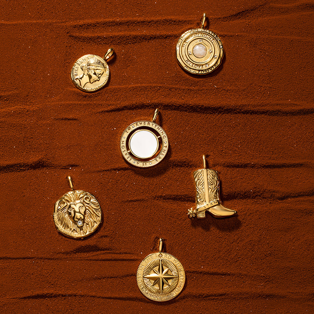 Five gold pendant charms arranged on a red dirt surface: a lion head pendant, a compass pendant, a cowboy boot pendant, a circular medallion pendant, and a circular pendant with a moonstone center.
