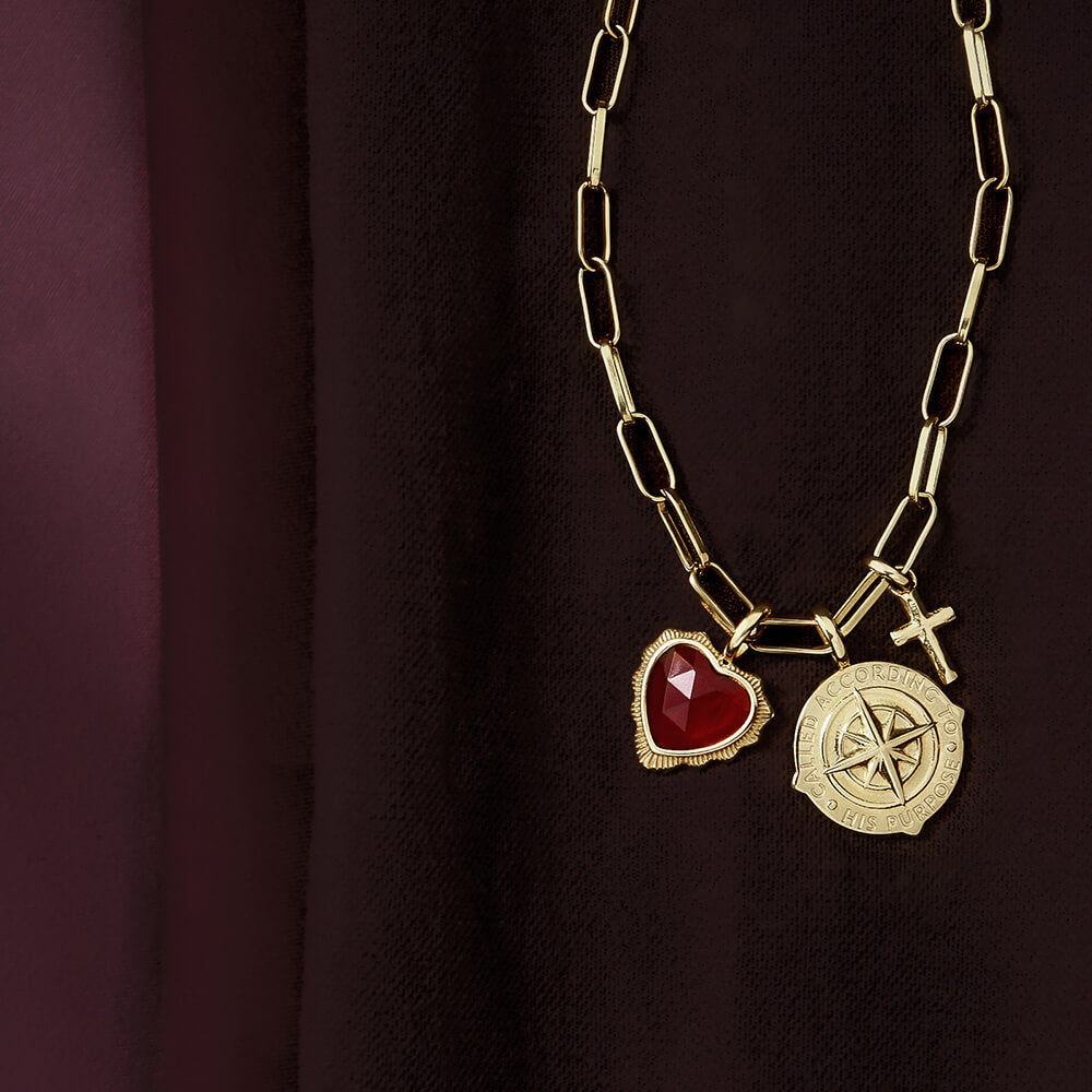 Gold necklace with red heart, cross and Called According to His Purpose medallion charms on burgundy fabric