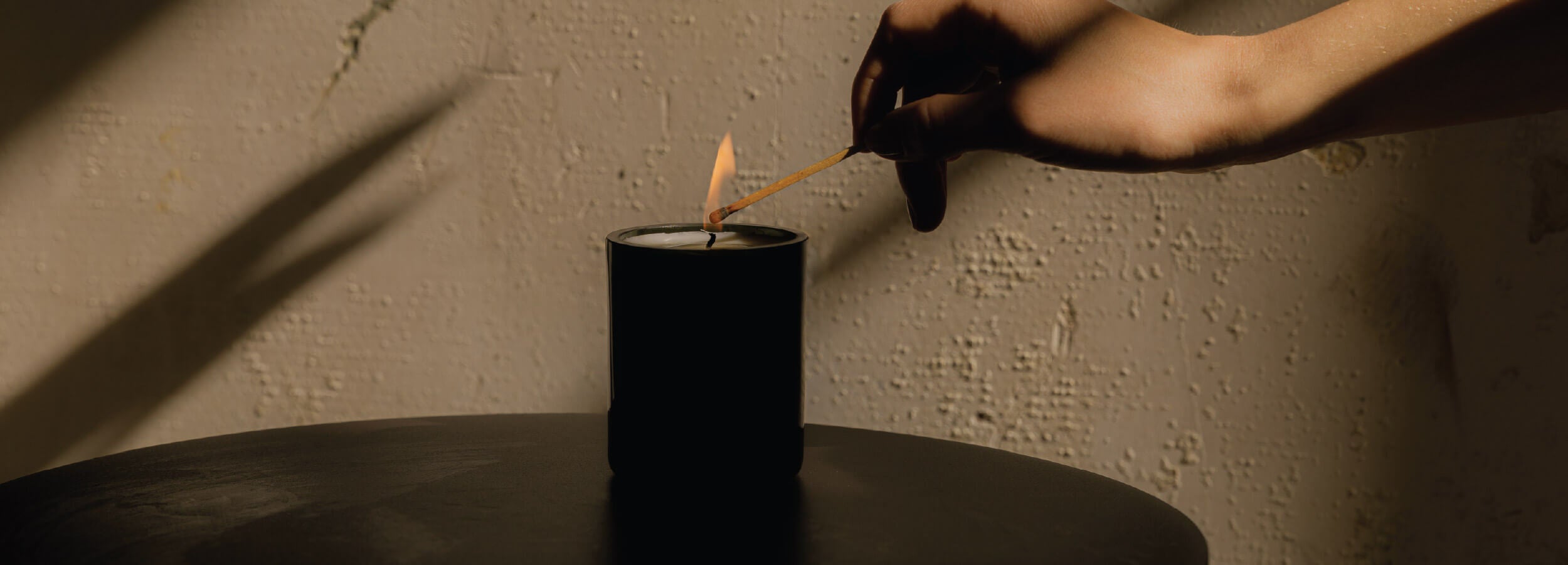 Person lighting a black candle with a match against a textured wall.