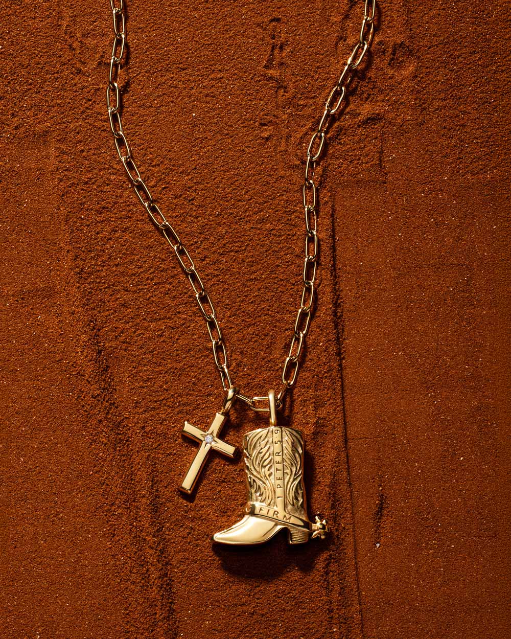 Gold cowboy boot and cross charms on a paperclip chain against textured rust-coloured sand.