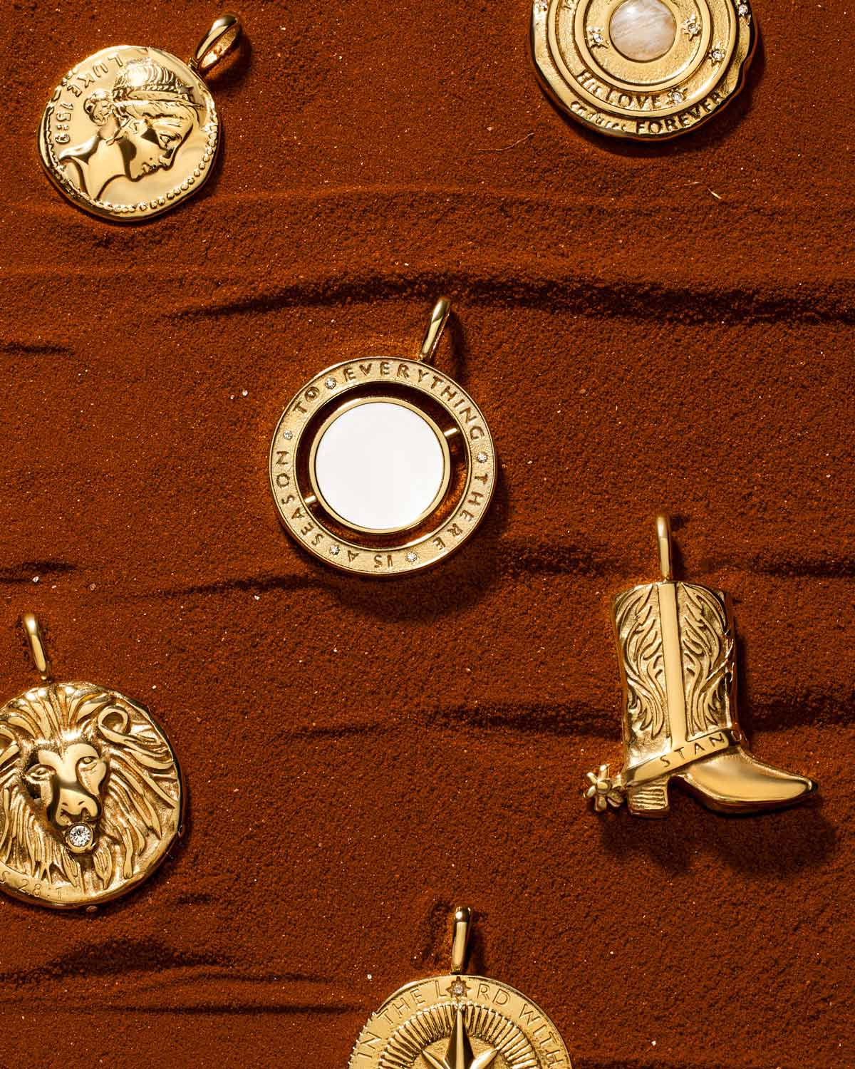 Six gold Christian medallions, including lion, cowboy boot, compass and scripture designs, arranged on sand