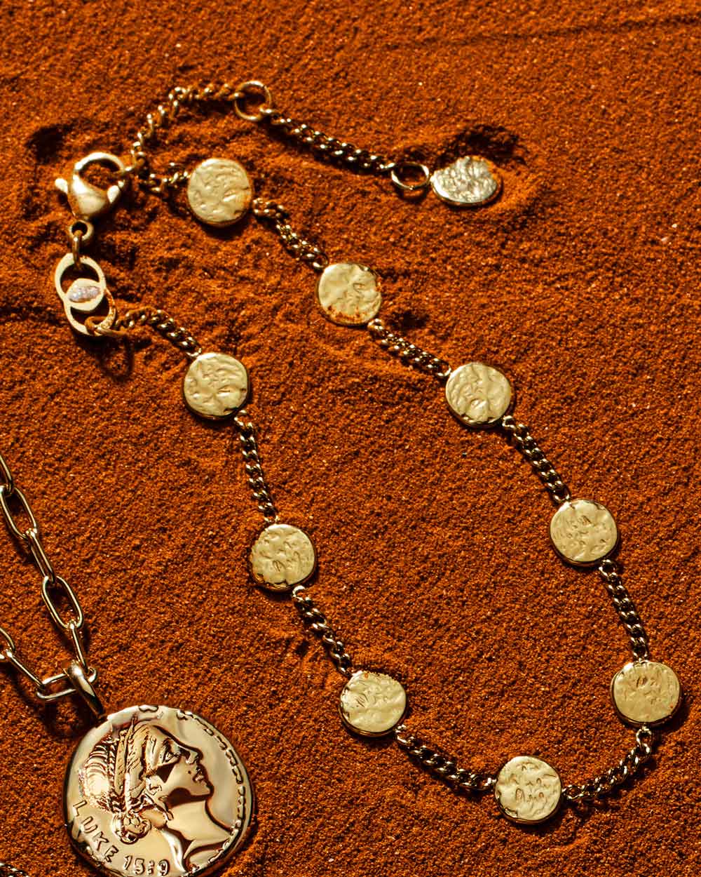 Gold Luke 15:9 coin pendant necklace and matching coin bracelet arranged on textured rust-coloured sand.